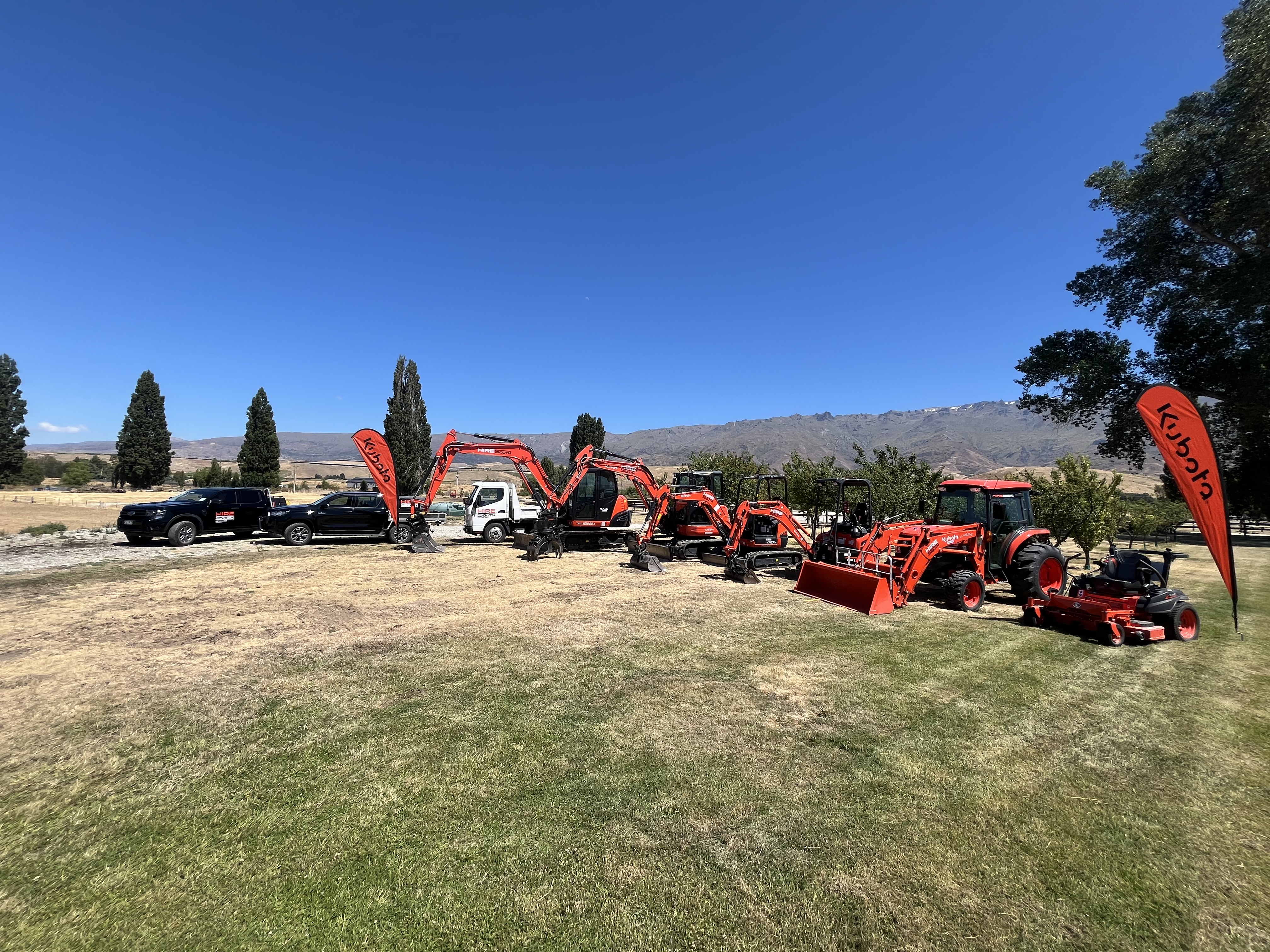 The Hire South fleet — multiple Kubota diggers, tractor, and support vehicles lined up with the Central Otago mountain ranges behind them