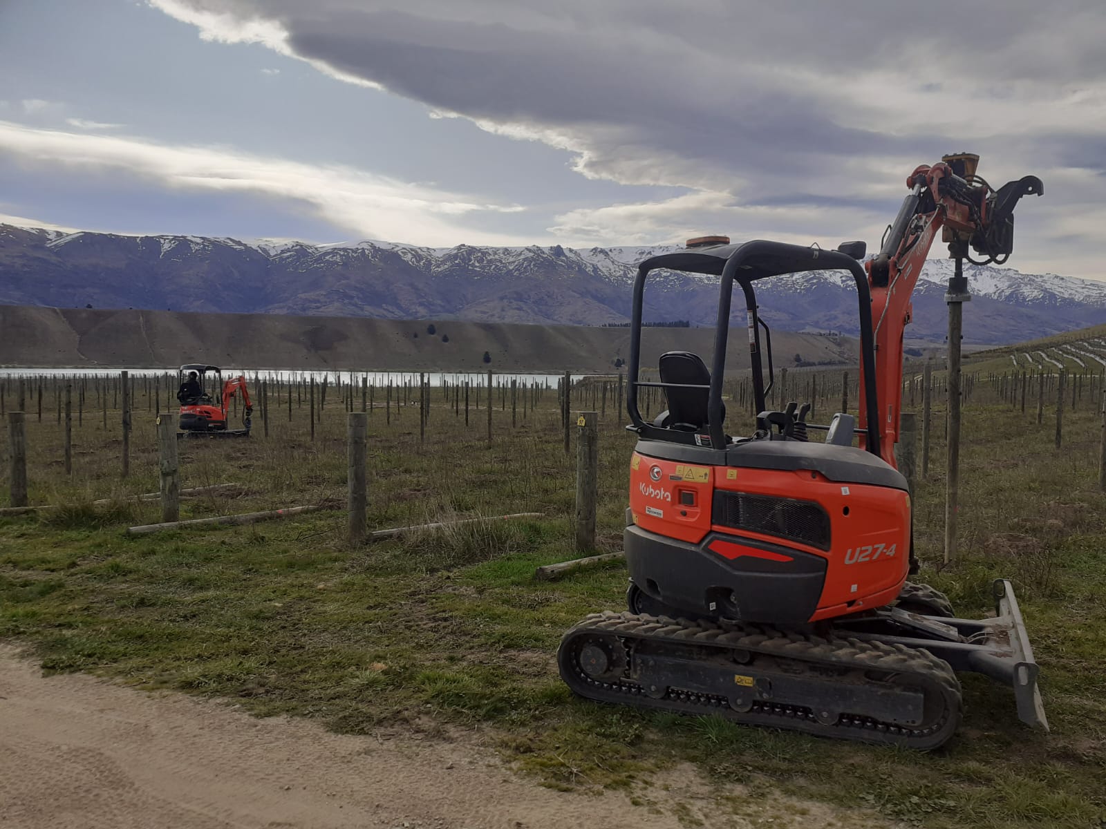 Hire South Kubota U27-4 digger parked in a vineyard row with Lake Dunstan and snowy Central Otago mountains visible under dramatic cloudy skies