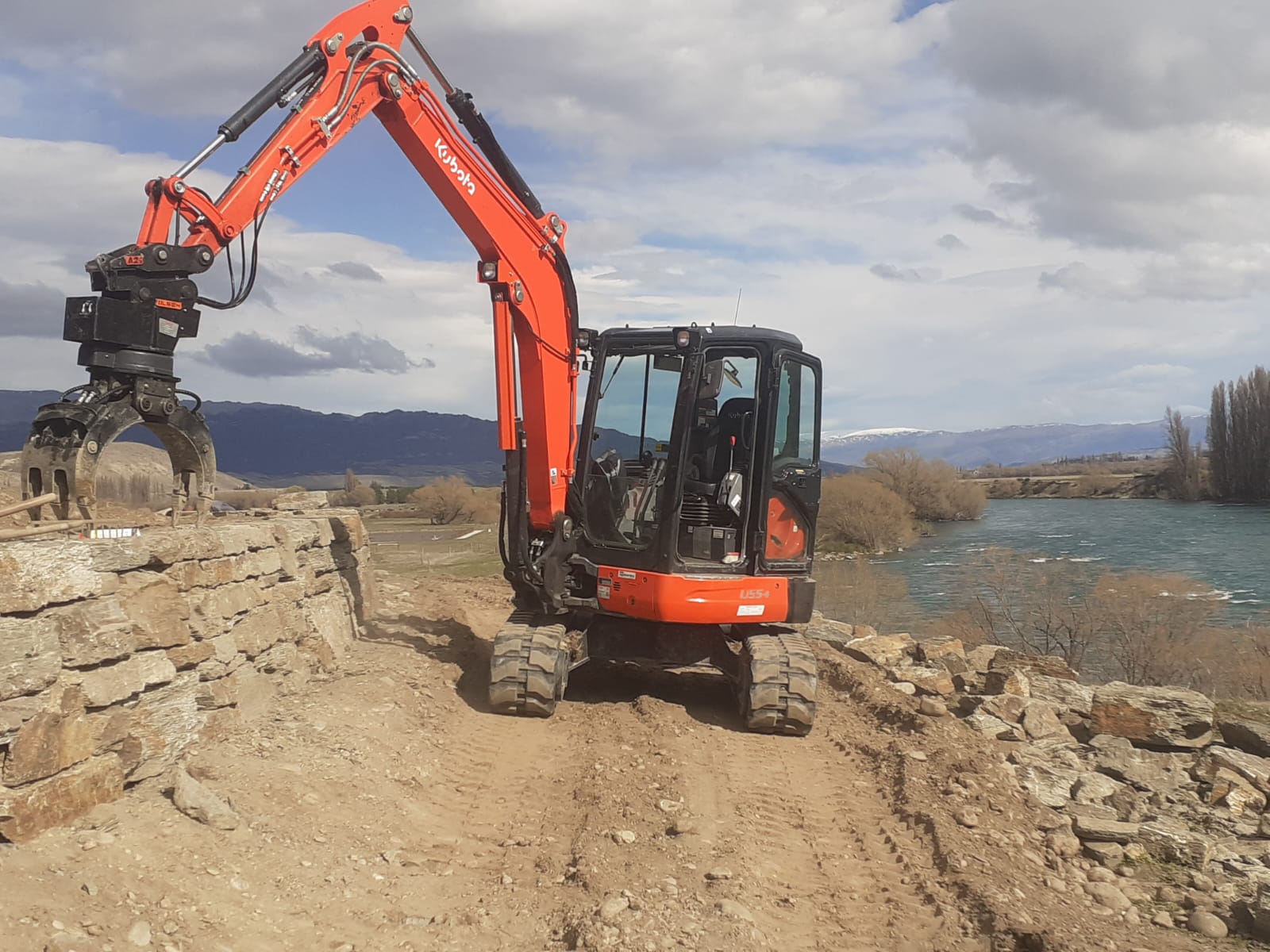 Hire South Kubota U55-4 digger fitted with a grapple attachment, working on a historic stone riverbank beside the Clutha River, Central Otago