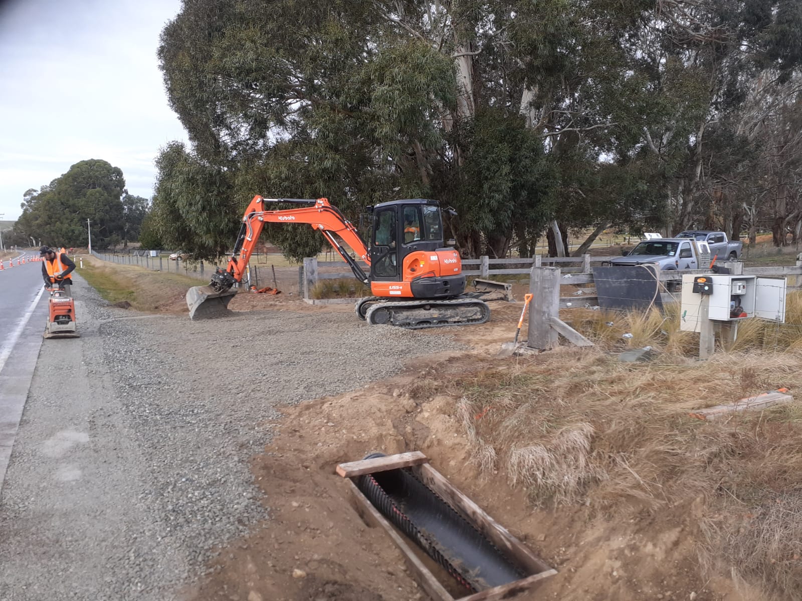 Hire South Kubota U55-4 digger and a worker using a plate compactor on a roadside civil construction job, gum trees lining the rural road