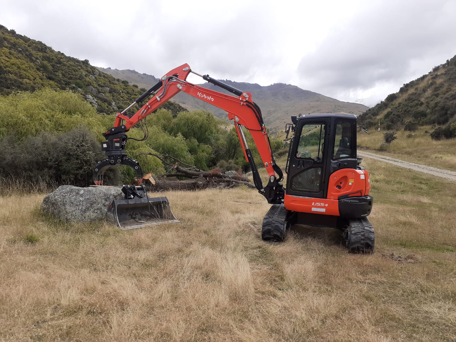 Hire South Kubota U55-4 digger with grapple and bucket attachments clearing scrub and logs in a rocky Central Otago gorge