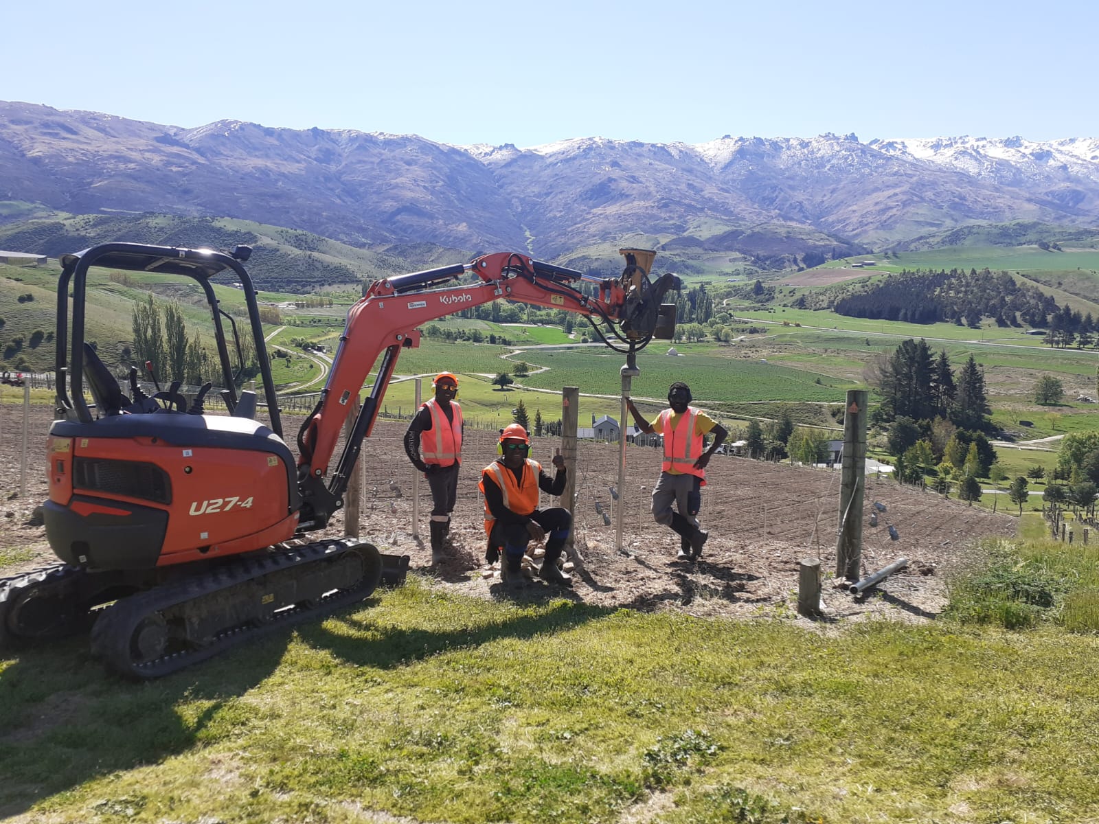 Hire South team of three workers in hi-vis with a Kubota U27-4 digger after completing a vineyard fence post driving job, Cromwell Basin panorama and snowy mountains behind