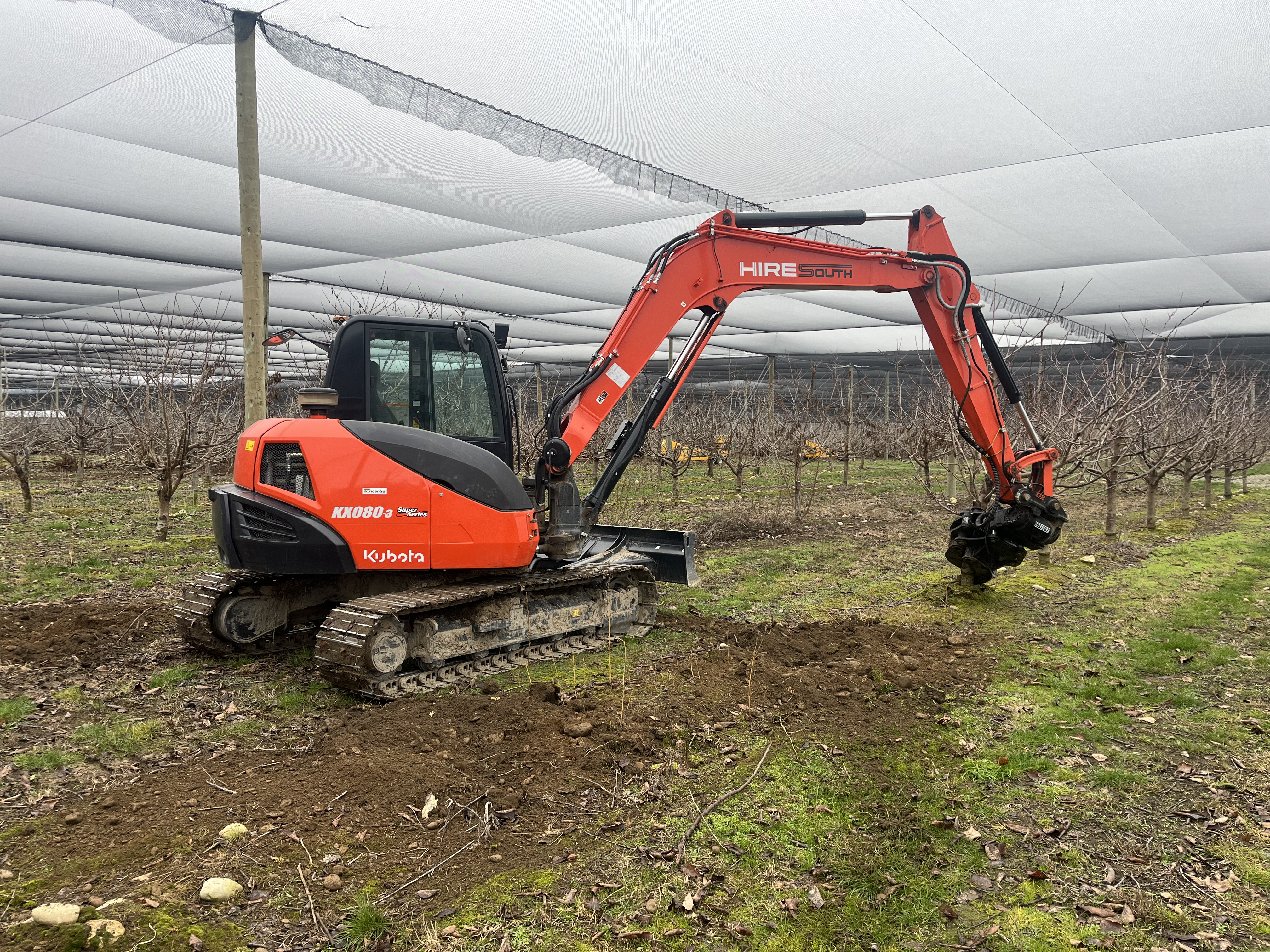 Hire South Kubota KX080-3 digger with grab attachment, side profile view working beneath stonefruit orchard shade netting, Central Otago