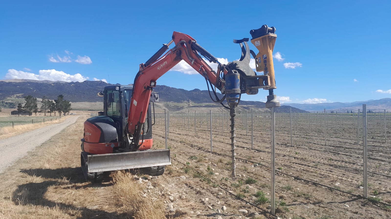 Kubota U55 digger fitted with a post driver attachment, working a vineyard row in Central Otago