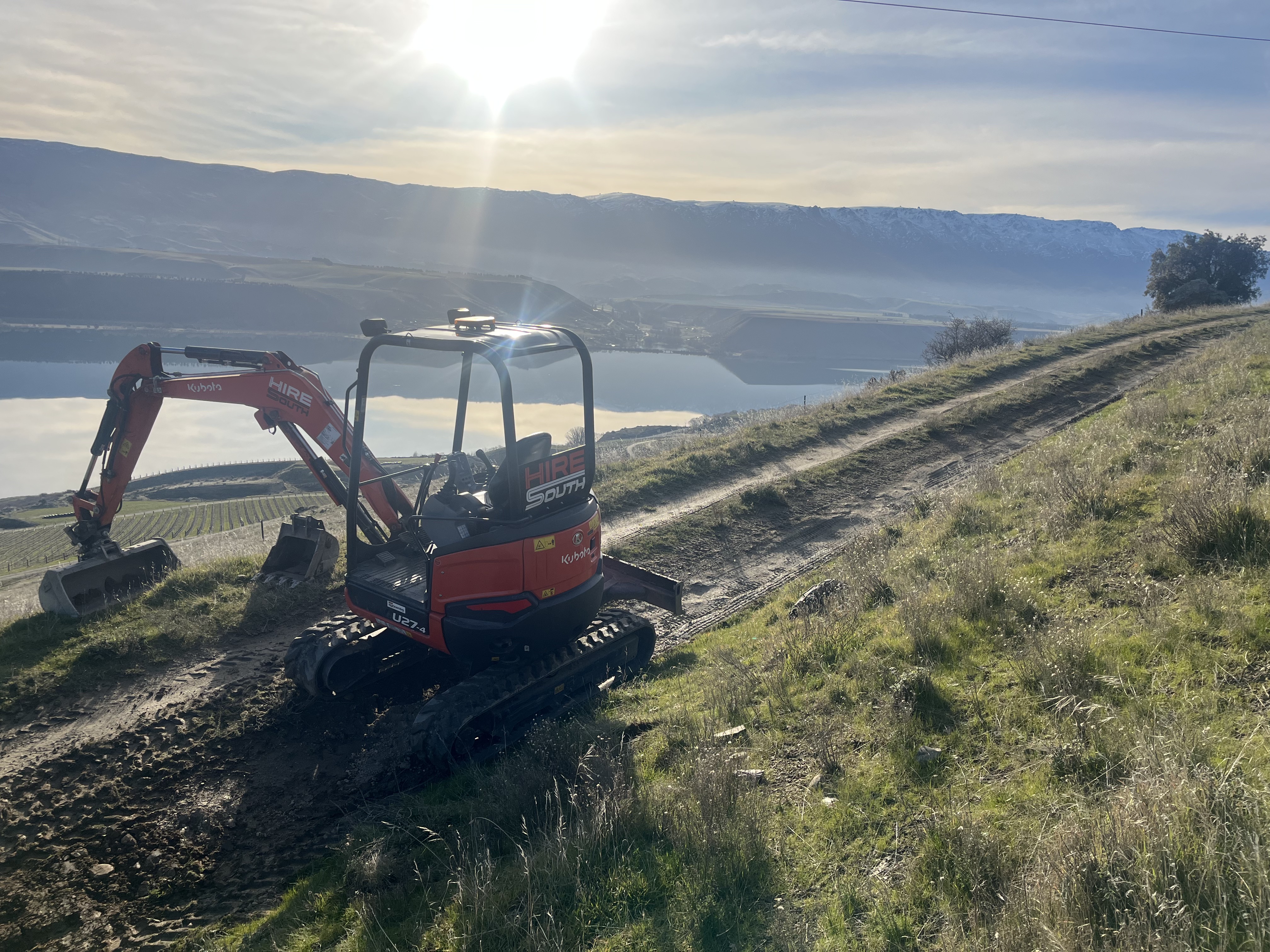 Hire South branded Kubota U27-4 digger working on a steep slope above Lake Dunstan, dramatic sunrise light with snow-capped mountains in the background, Cromwell