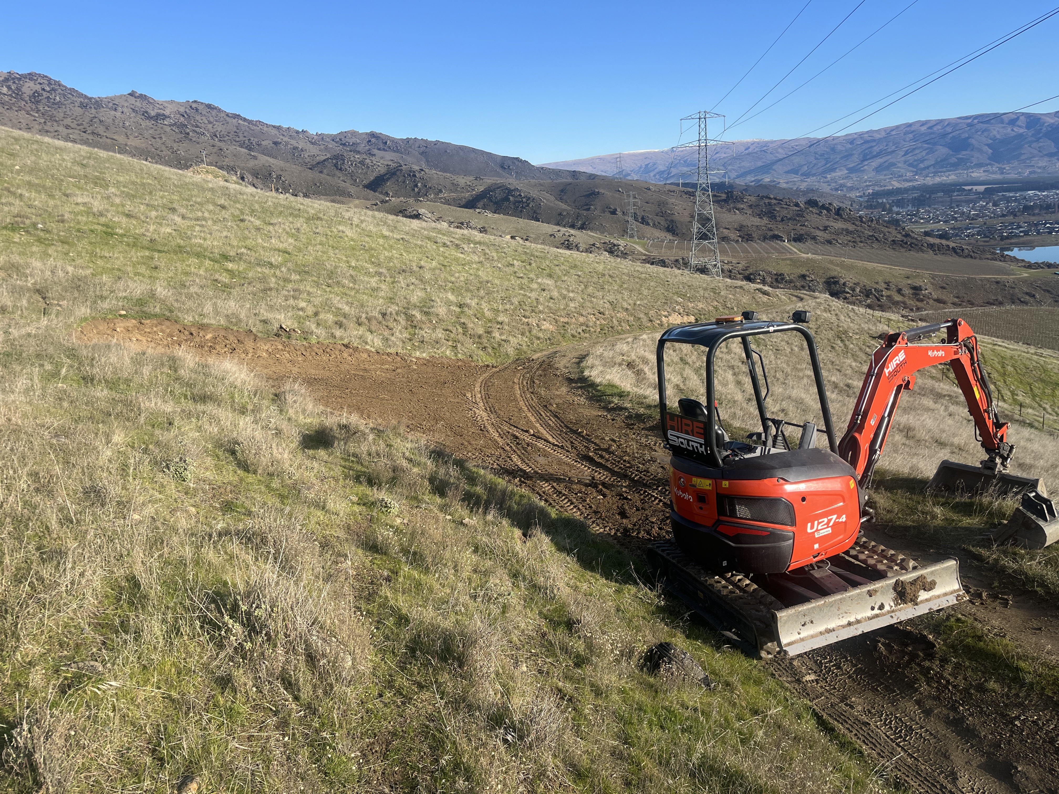 Hire South Kubota U27-4 digger forming a new access track on a steep tussock hillside above Cromwell and Lake Dunstan, Central Otago — high-voltage transmission lines and mountains behind